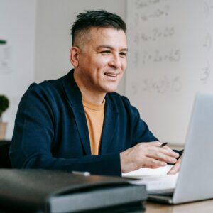 A person smiling while working on a laptop with written notes and a pen, in a modern classroom setting.
