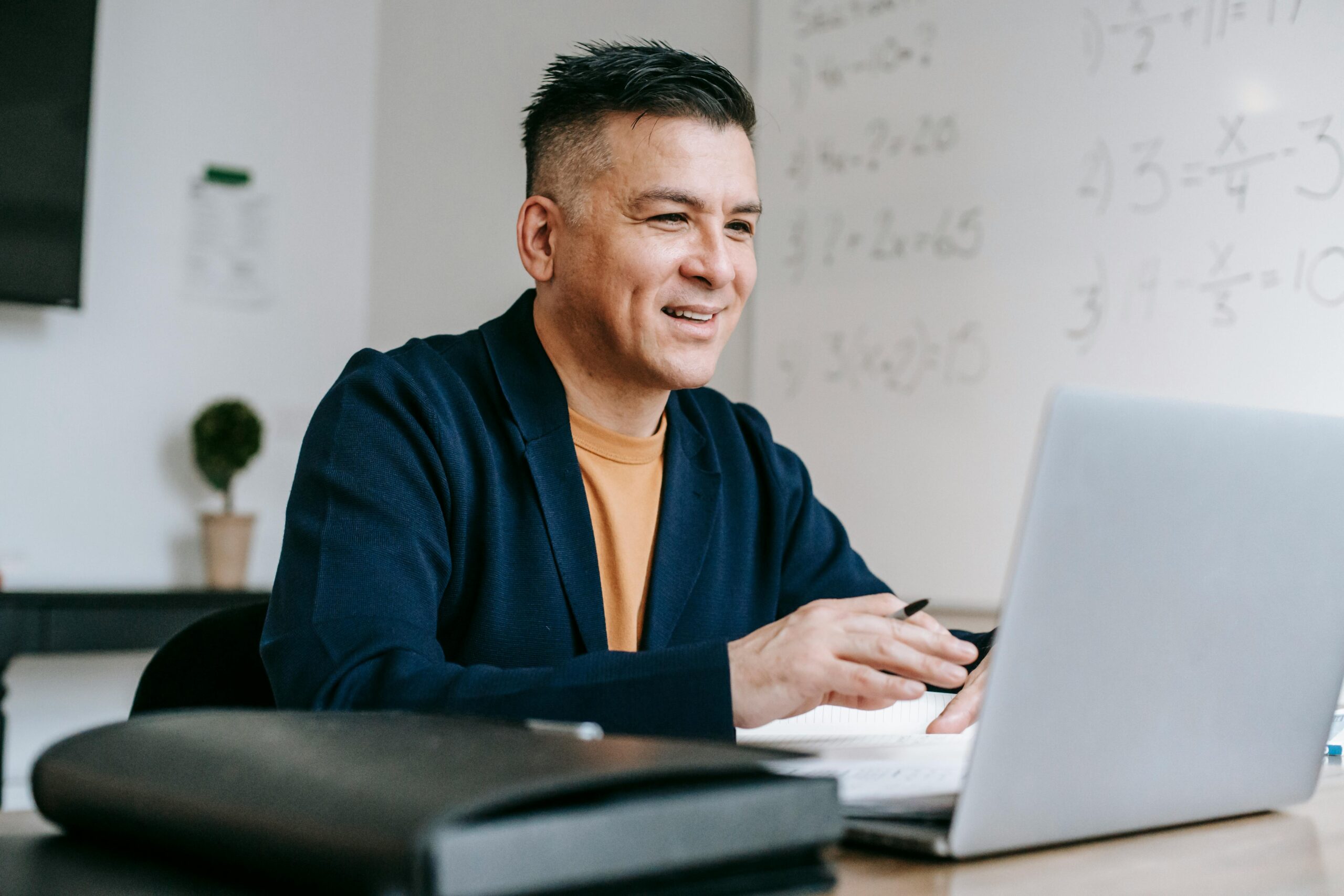 A person smiling while working on a laptop with written notes and a pen, in a modern classroom setting.