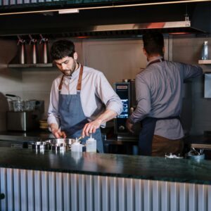 Two chefs working in a modern kitchen, one at a stovetop and another using an oven, preparing dishes.