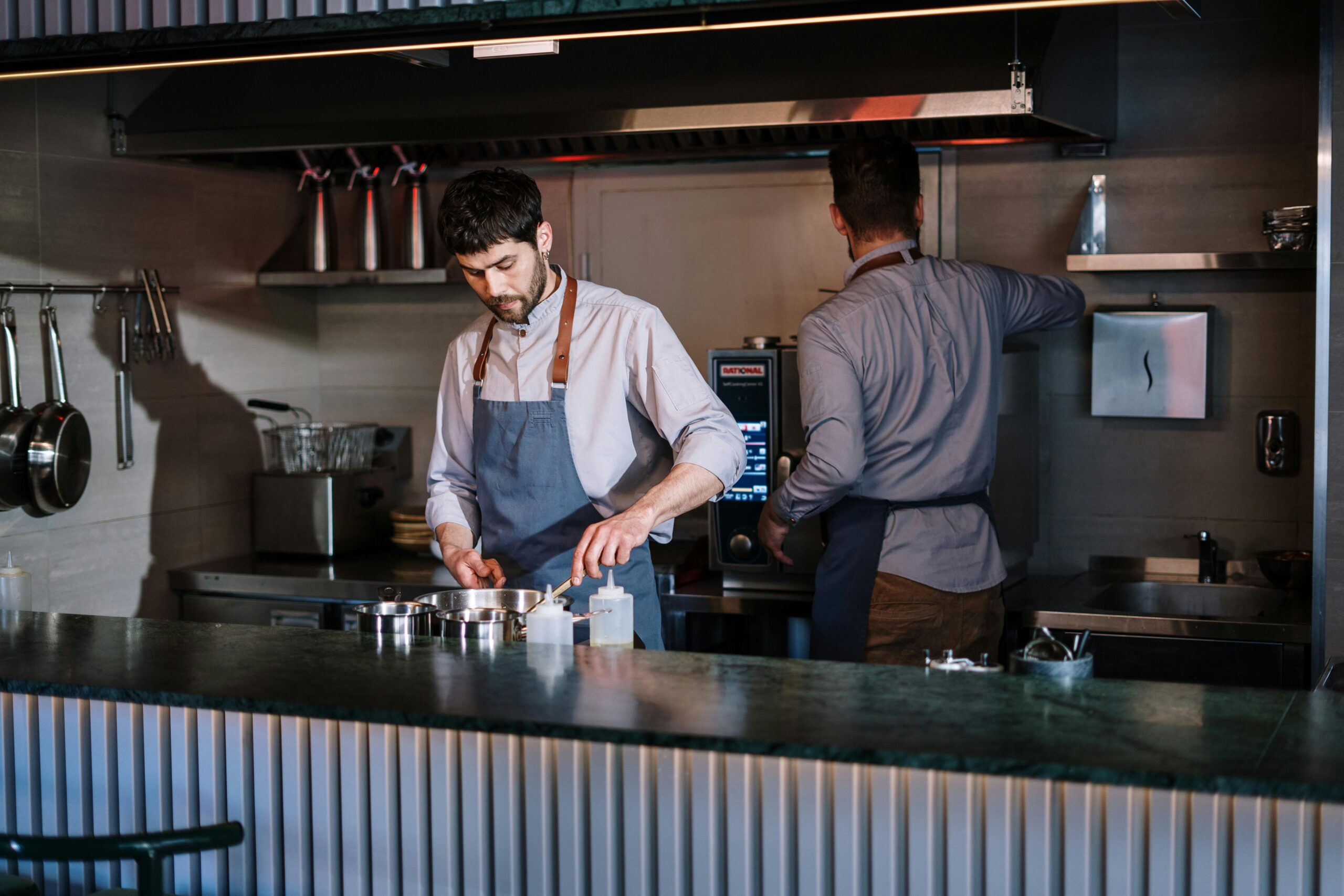 Two chefs working in a modern kitchen, one at a stovetop and another using an oven, preparing dishes.