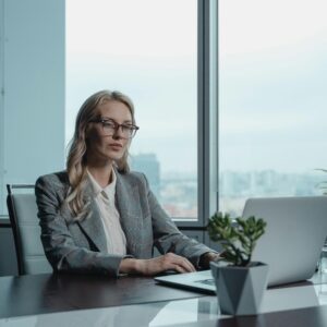 A person in a suit seated at a desk with a laptop, in an office with large windows and plants.