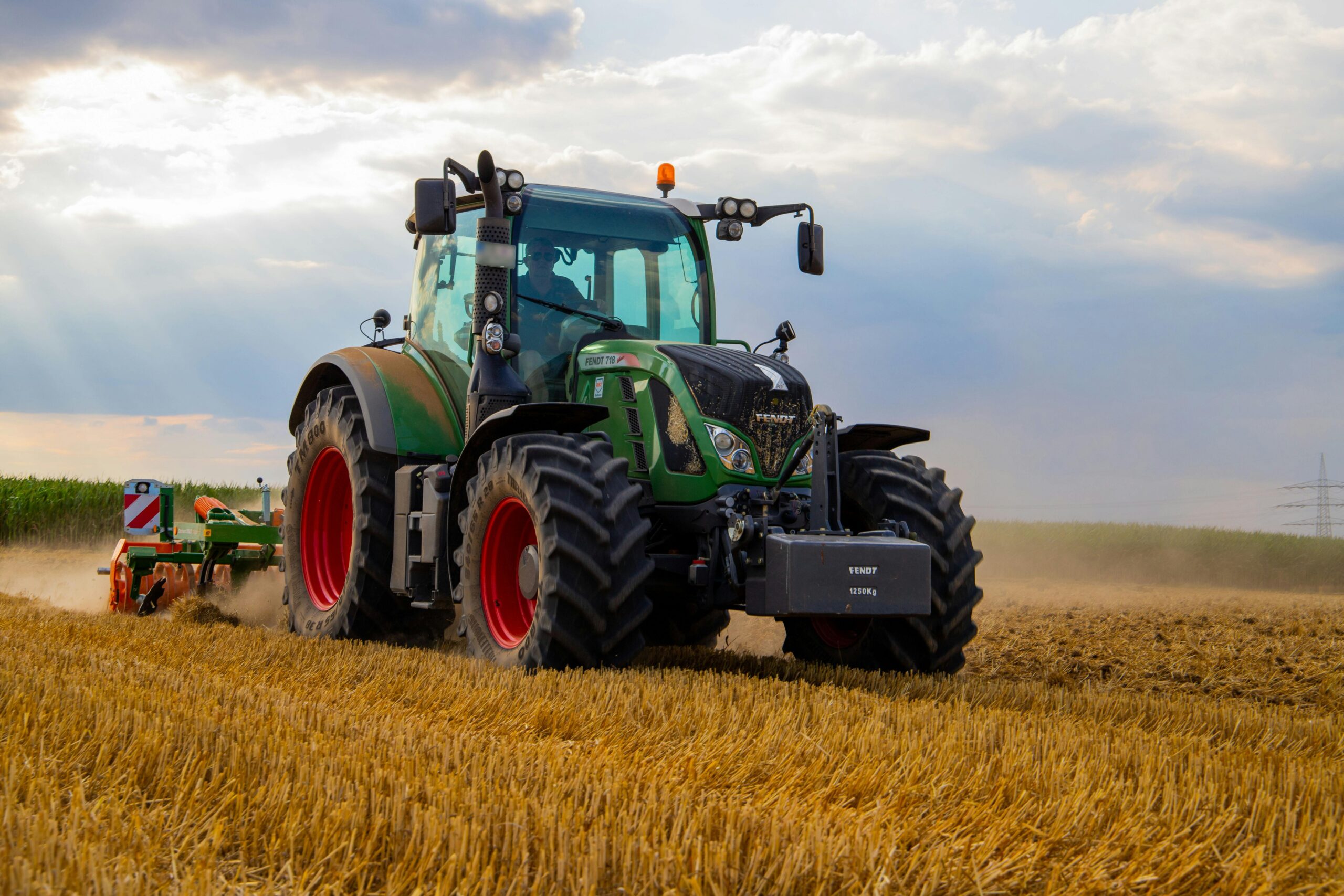 A person drives a green tractor plowing a field under a cloudy sky.
