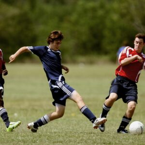 Several athletes competing in a soccer match on a grassy field, two in red jerseys, one in blue, engaged in action.