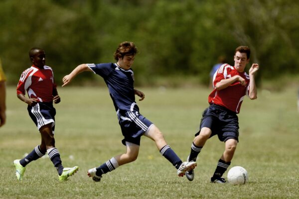 Several athletes competing in a soccer match on a grassy field, two in red jerseys, one in blue, engaged in action.