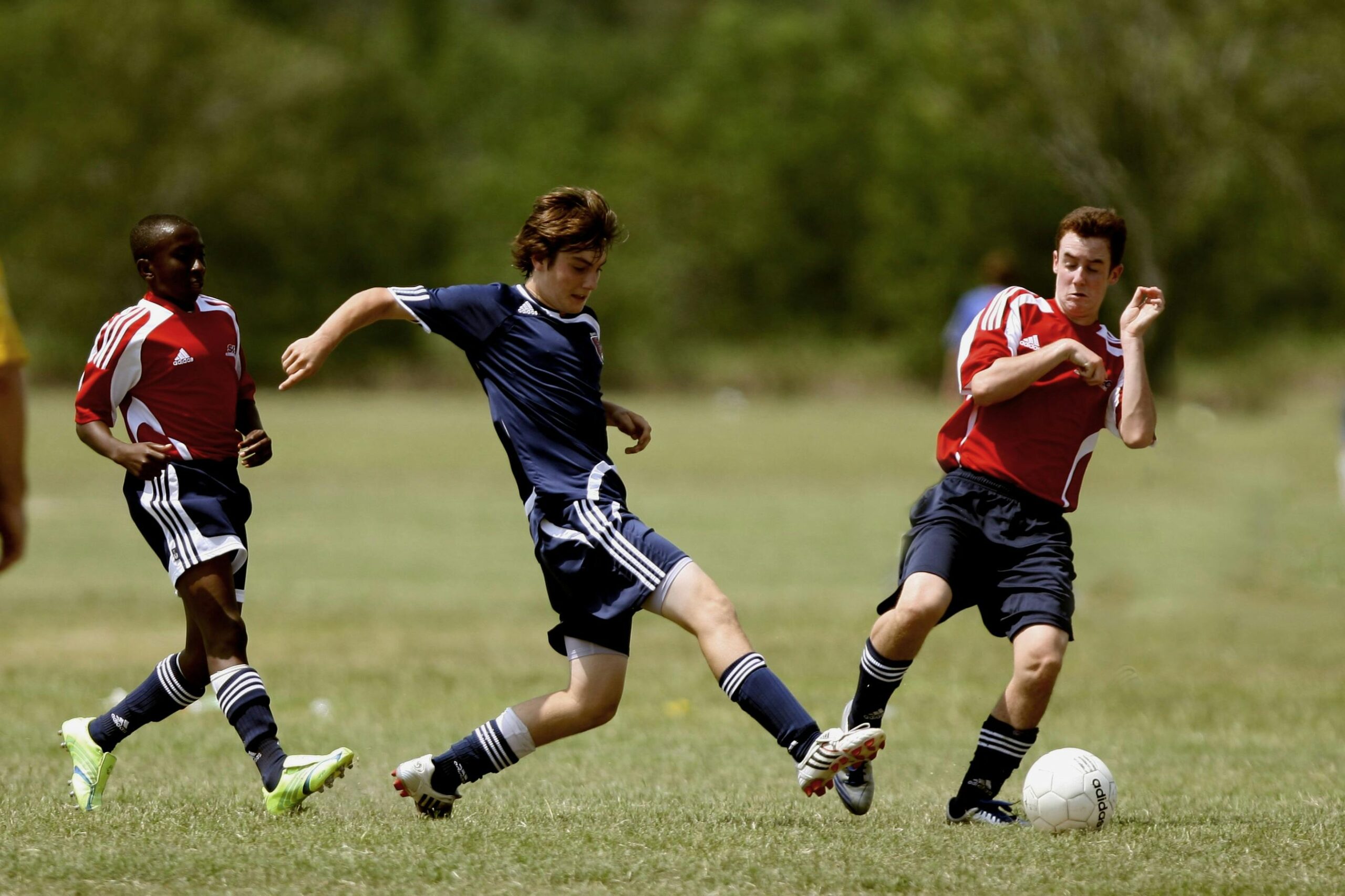 Several athletes competing in a soccer match on a grassy field, two in red jerseys, one in blue, engaged in action.