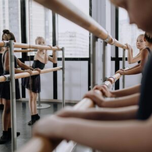 Several students practicing ballet at a barre in a dance studio with large windows.