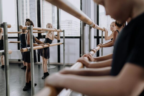 Several students practicing ballet at a barre in a dance studio with large windows.