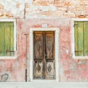 Weathered red wall with green window shutters and an aged wooden door, reflecting rustic charm and vibrant textures.