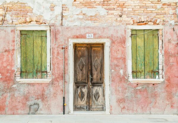 Weathered red wall with green window shutters and an aged wooden door, reflecting rustic charm and vibrant textures.