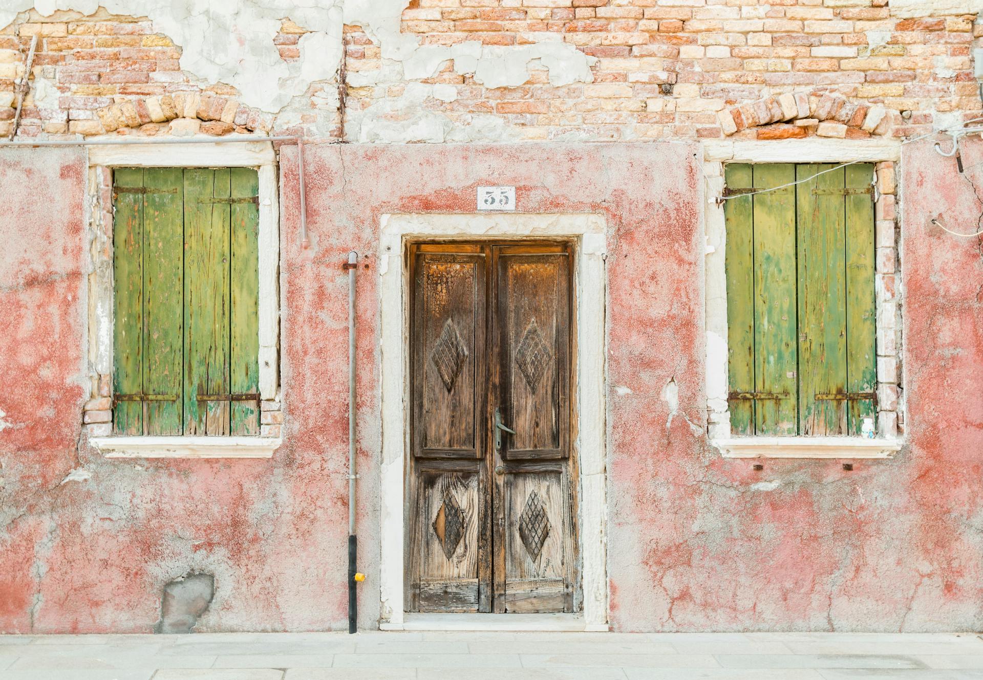Weathered red wall with green window shutters and an aged wooden door, reflecting rustic charm and vibrant textures.