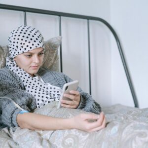 A person with a headscarf lies in bed using a smartphone, with an IV line in their arm.