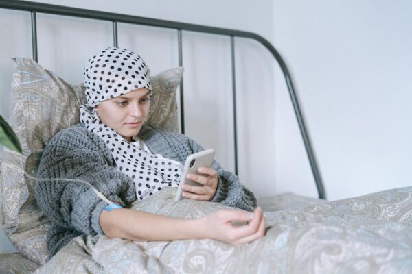 A person with a headscarf lies in bed using a smartphone, with an IV line in their arm.