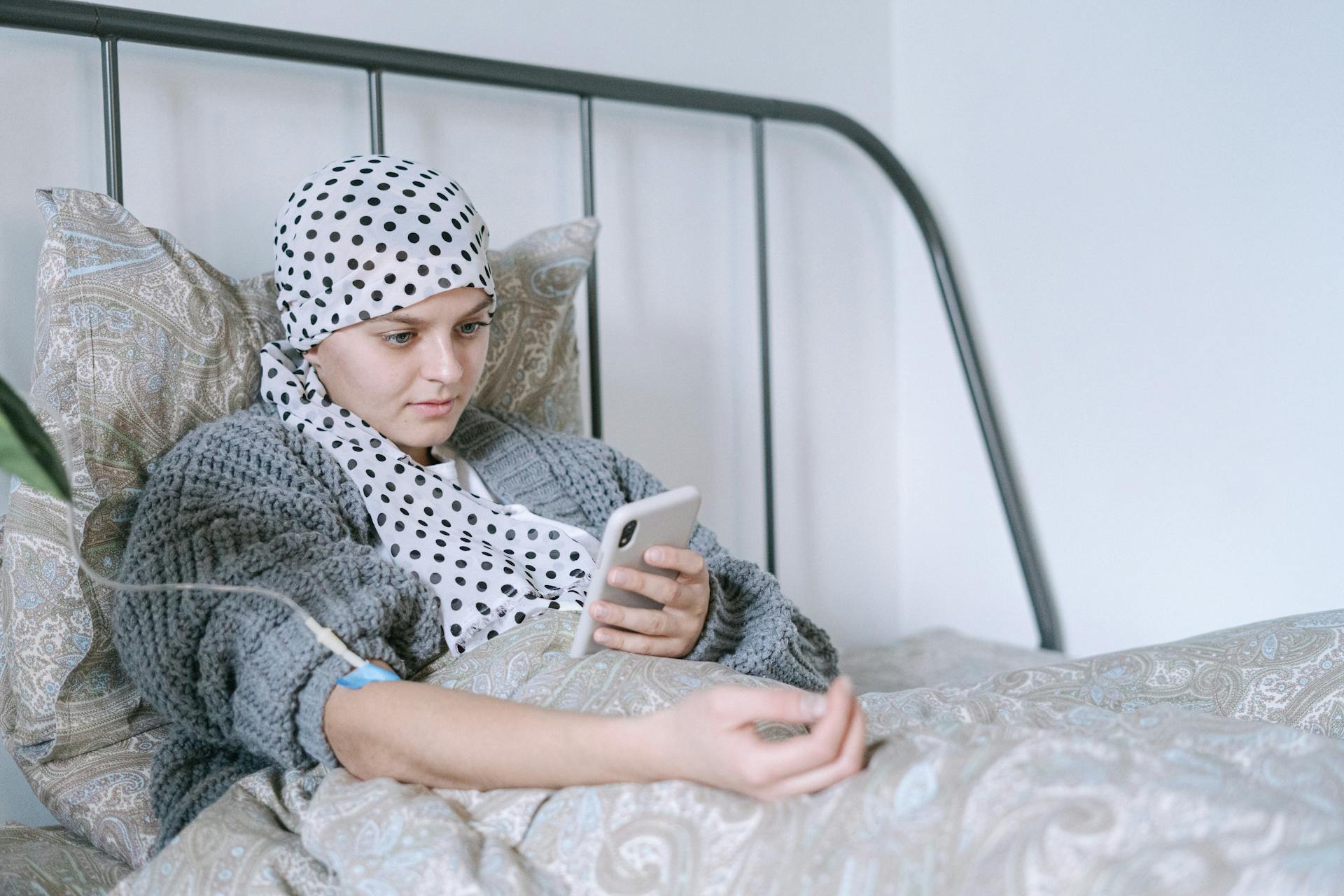 A person with a headscarf lies in bed using a smartphone, with an IV line in their arm.