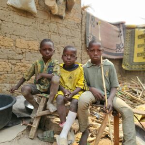 Three children sitting on benches outside by a brick wall, surrounded by various household items and rugs.