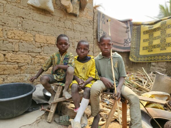 Three children sitting on benches outside by a brick wall, surrounded by various household items and rugs.