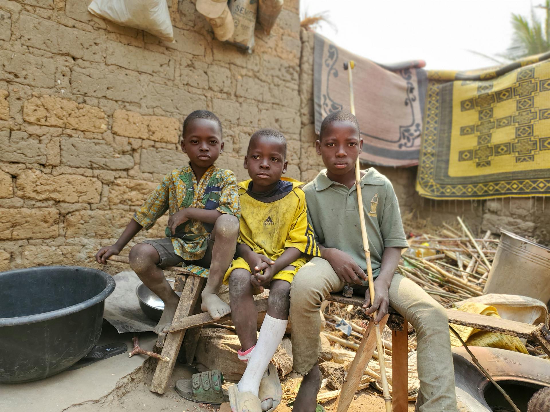 Three children sitting on benches outside by a brick wall, surrounded by various household items and rugs.