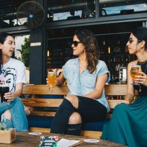 Three people sitting on a bench outside, enjoying drinks and talking casually.