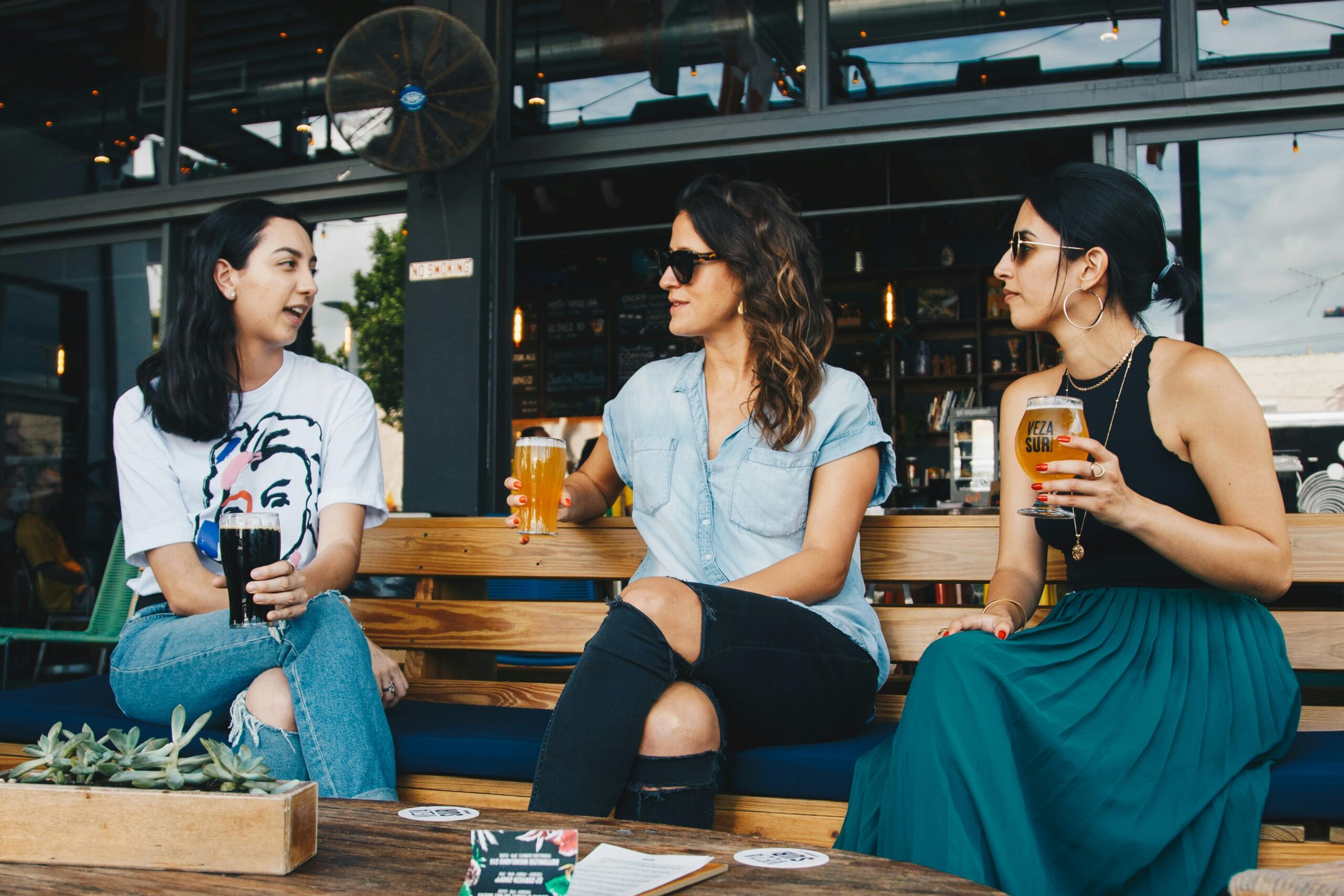 Three people sitting on a bench outside, enjoying drinks and talking casually.
