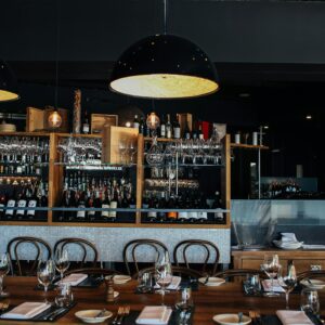 Cozy restaurant interior with wooden tables, wine bottles, and glasses, illuminated by modern pendant lights.