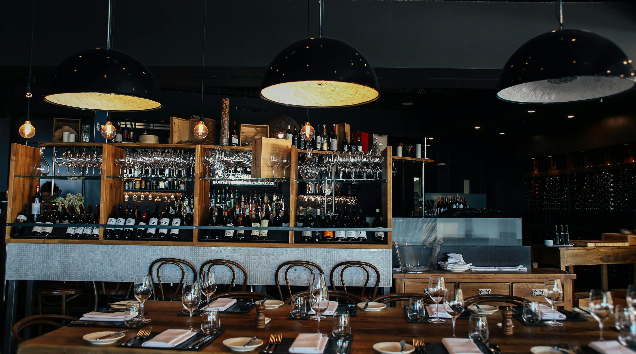 Cozy restaurant interior with wooden tables, wine bottles, and glasses, illuminated by modern pendant lights.