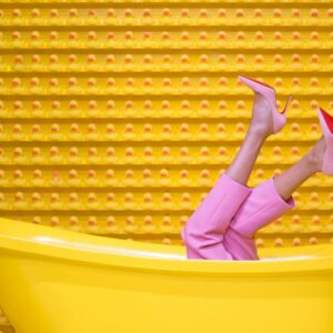 Legs in pink pants and high heels sticking out of a yellow tub, with a wall of rubber ducks in the background.