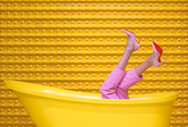 Legs in pink pants and high heels sticking out of a yellow tub, with a wall of rubber ducks in the background.