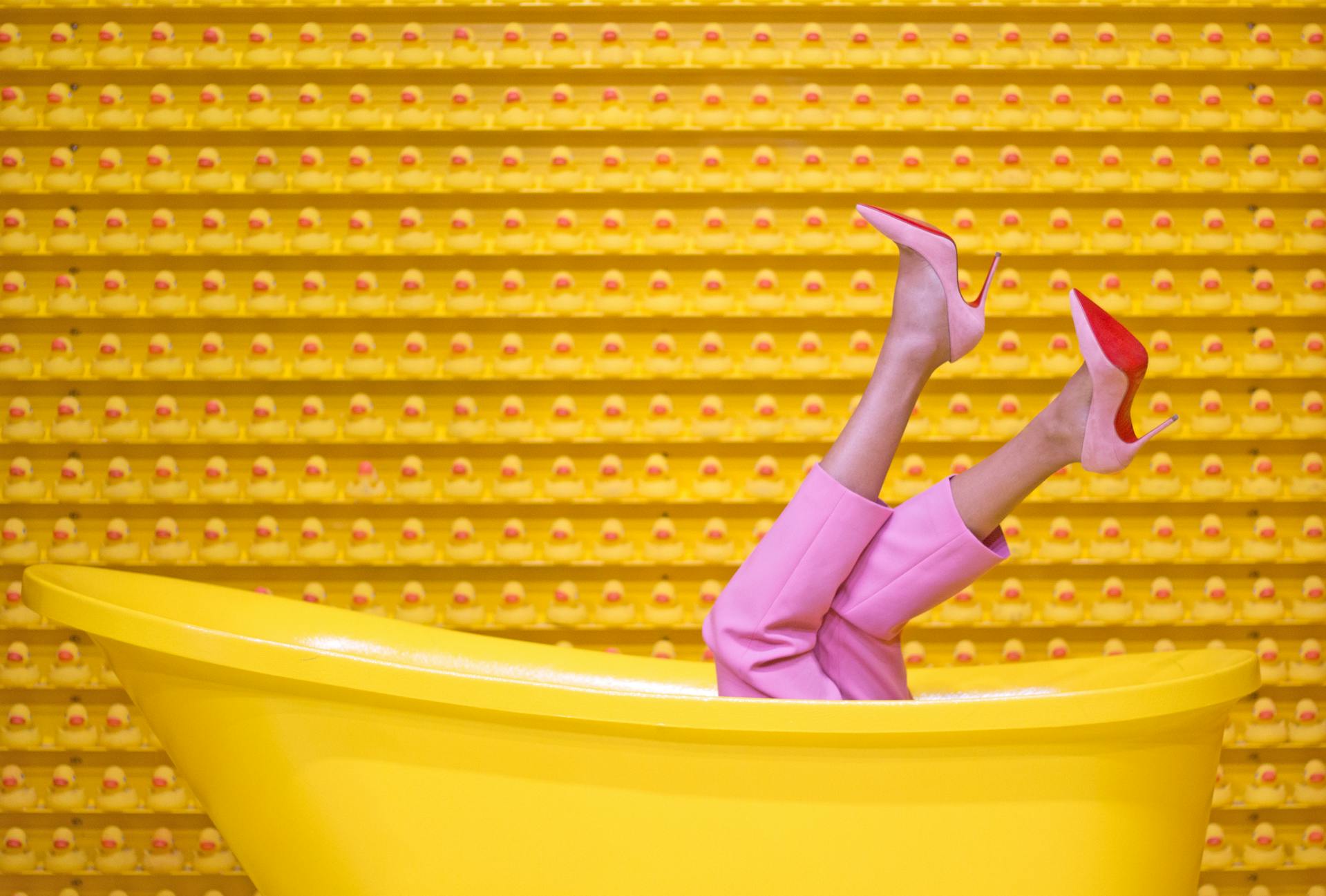 Legs in pink pants and high heels sticking out of a yellow tub, with a wall of rubber ducks in the background.