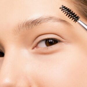 A person using an eyebrow brush to style their eyebrow against a light blue background.