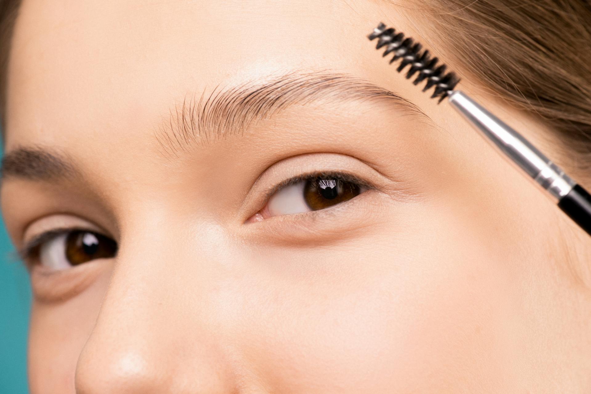 A person using an eyebrow brush to style their eyebrow against a light blue background.