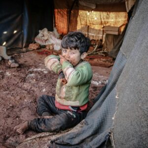 A child sitting on muddy ground inside a dimly lit tent, wearing a green sweater.