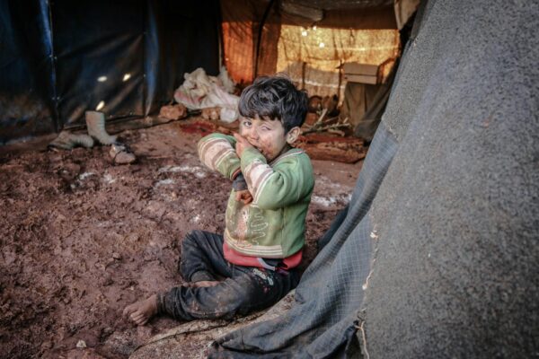 A child sitting on muddy ground inside a dimly lit tent, wearing a green sweater.