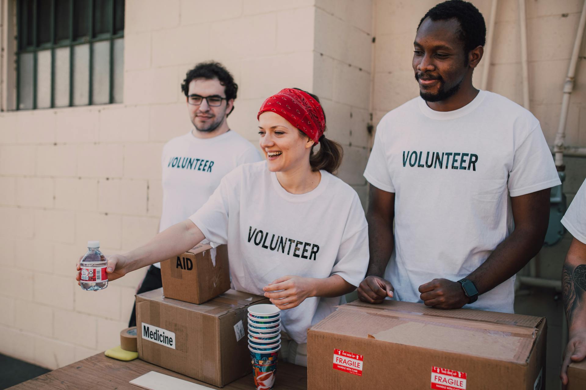 Volunteers distributing supplies, smiling and organizing boxes labeled aid and medicine on a table outdoors.