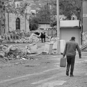 A person carrying a container walks through a street with debris and damaged buildings in the background.
