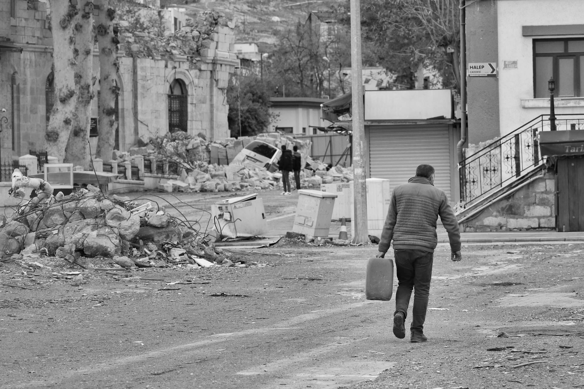 A person carrying a container walks through a street with debris and damaged buildings in the background.