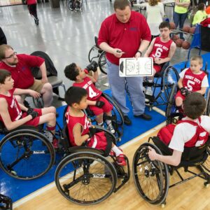 A coach discusses strategies with a wheelchair basketball team wearing red jerseys in a gym.