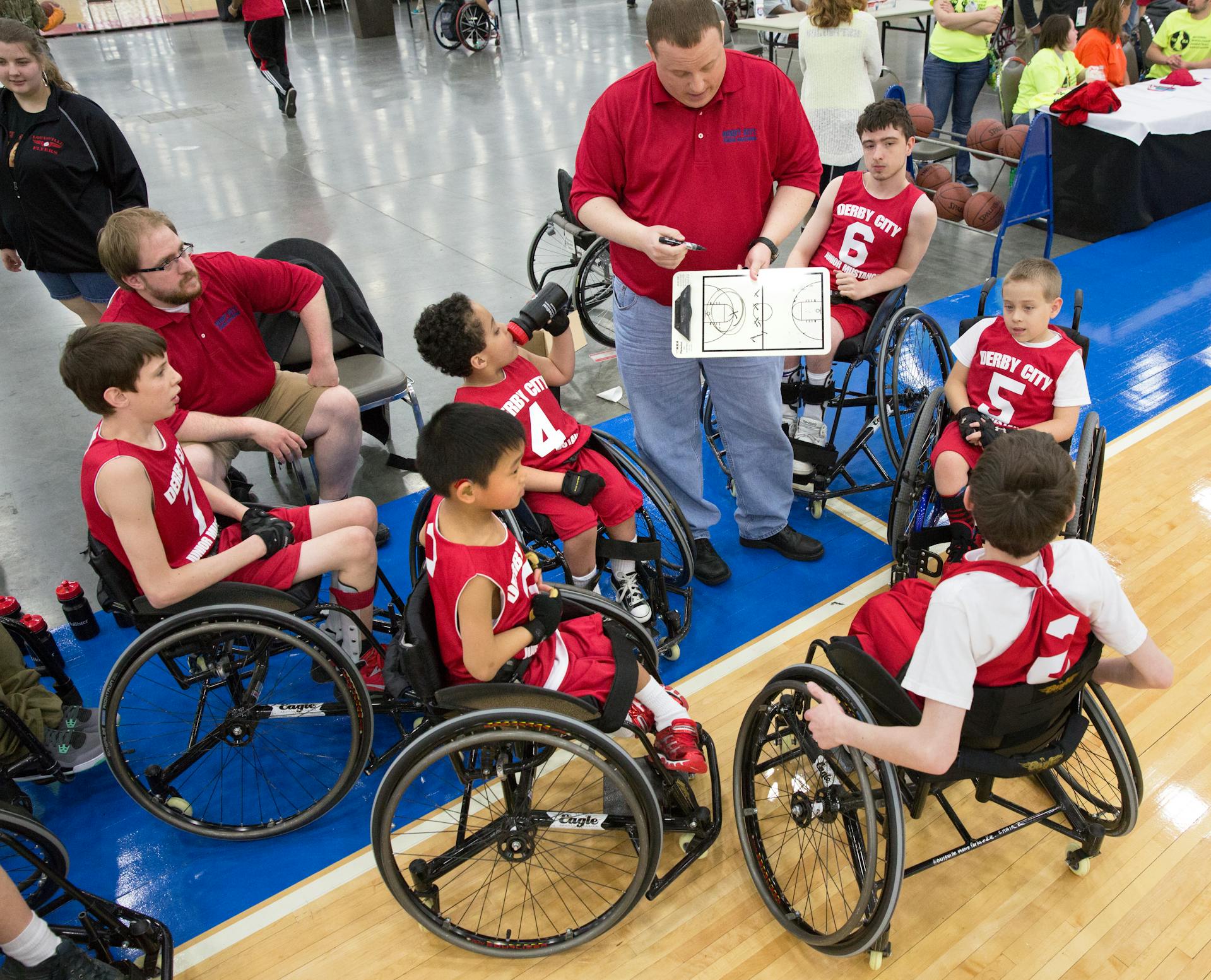 A coach discusses strategies with a wheelchair basketball team wearing red jerseys in a gym.