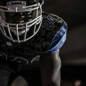 A person in a football uniform and helmet, crouched and ready to play, on a dark background.