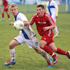 Two soccer players in action on a grassy field, one in red and the other in white, competing for the ball.