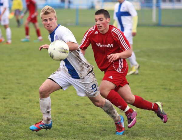 Two soccer players in action on a grassy field, one in red and the other in white, competing for the ball.