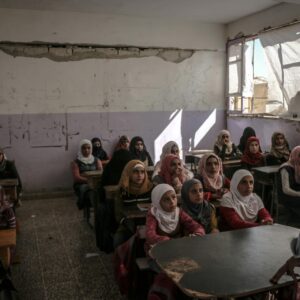 A classroom with several students sitting at desks, listening to a person speaking in the front.