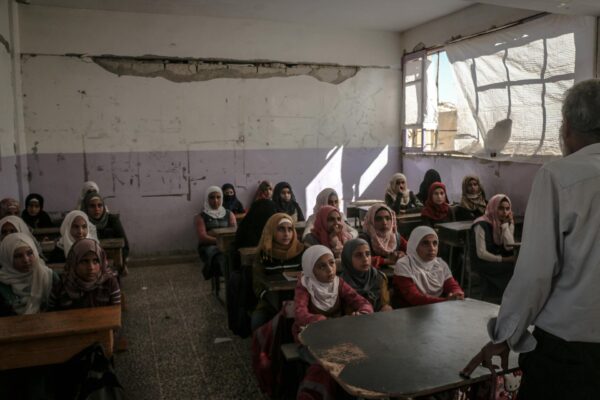 A classroom with several students sitting at desks, listening to a person speaking in the front.